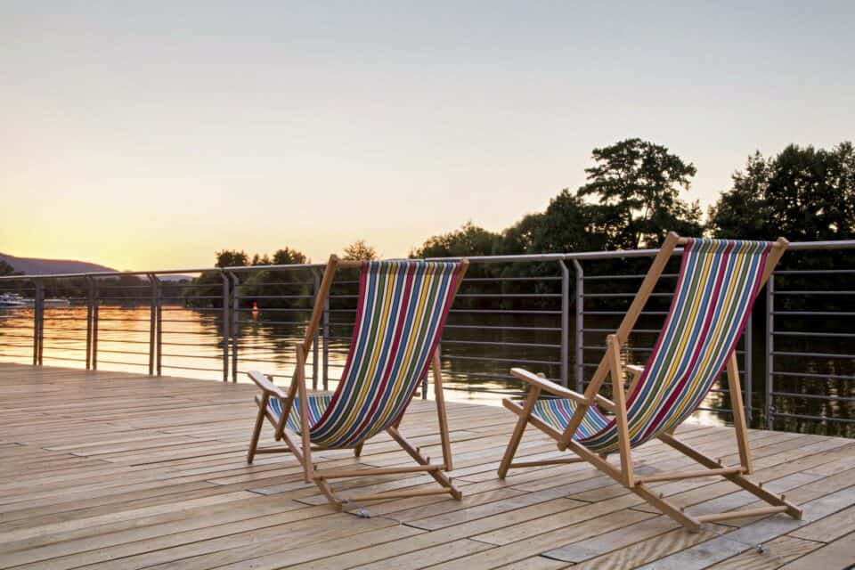 MainZauber Apartments früher MainTerrasse Miltenberg am Main - Entdecken Sie die perfekte Ferienwohnung mit atemberaubender Aussicht! Stellen Sie sich vor, Sie entspannen sich in einer luxuriösen Unterkunft auf zwei bequemen Liegestühlen auf einer Holzterrasse mit Blick auf einen ruhigen See bei Sonnenuntergang. Erleben Sie den atemberaubenden Himmel, der in warmen Farbtönen leuchtet, während sich im Hintergrund majestätische Bäume abzeichnen. Diese Luxuswohnung bietet die Bühne für Ruhe und Entspannung und bietet Ihnen einen unvergesslichen Urlaub.