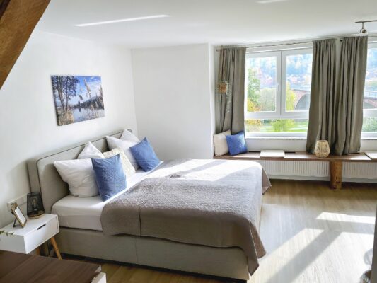 MainZauber Apartments früher MainTerrasse Miltenberg am Main - Entdecken Sie die perfekte Ferienwohnung mit einem Hauch von Luxus. Diese atemberaubende Luxuswohnung für den Urlaub verfügt über ein helles und einladendes Schlafzimmer mit einem geräumigen Bett mit eleganter grau-weißer Bettwäsche und flauschigen blauen Kissen. Genießen Sie das natürliche Sonnenlicht, das durch die mit eleganten grauen Vorhängen behangenen Fenster fällt. Tauchen Sie ein in die Ruhe dieses Raums, mit malerischen Kunstwerken, die den Wänden Charme verleihen, und einem gemütlichen Fenstersitz mit bequemen Kissen – ideal zum Entspannen nach Ihren täglichen Abenteuern. Wählen Sie für Ihren nächsten Urlaub eine unvergleichliche Unterkunft und gönnen Sie sich Komfort und Stil.