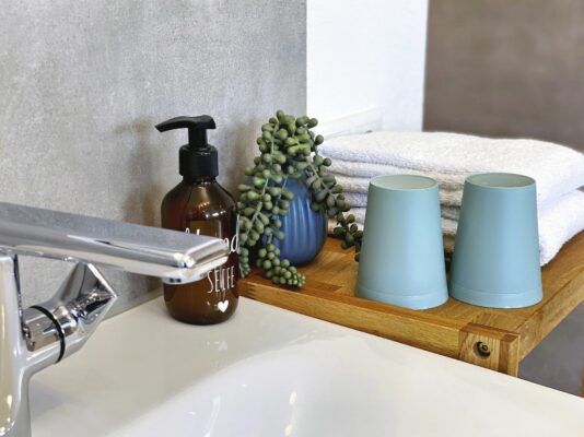 MainZauber Apartments früher MainTerrasse Miltenberg am Main - Entdecken Sie die perfekte Ferienwohnung mit einem luxuriösen Badezimmer, das moderne Eleganz und Komfort vereint. Erleben Sie die Eleganz eines modernen Wasserhahns und Waschbeckens, ergänzt durch wichtige Annehmlichkeiten wie einen stilvollen Seifenspender und üppiges Grün für eine erfrischende Note. Genießen Sie flauschige weiße Handtücher, die ordentlich auf einem schicken Holzregal gestapelt sind, neben zwei klassischen blauen Tassen – ideal, um Ihre Unterkunft noch komfortabler zu gestalten. Genießen Sie jeden Moment Ihres Luxusapartments für den Urlaub, wo Stil auf Ruhe trifft.