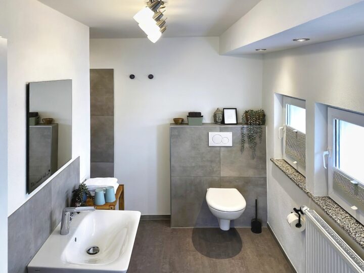MainZauber Apartments früher MainTerrasse Miltenberg am Main - Erleben Sie das ultimative Maß an Entspannung und Stil in dieser luxuriösen Ferienwohnung mit elegantem, modernem Badezimmer. Diese Unterkunft verfügt über eine elegante Wandtoilette und ein Waschbecken, ergänzt durch schicke graue Fliesen, die sowohl die Wände als auch den Boden bedecken. Verschönern Sie Ihren Aufenthalt in dieser ruhigen Umgebung mit zwei bezaubernden Pflanzen, die den Raum betonen. Genießen Sie reichlich natürliches Licht, das durch drei einladende Fenster auf der rechten Seite einfällt und eine helle und luftige Atmosphäre schafft. Verwöhnen Sie sich mit flauschigen Handtüchern, die sorgfältig neben dem Waschbecken angeordnet sind und Ihrem perfekten Urlaubsdomizil eine luxuriöse Note verleihen.