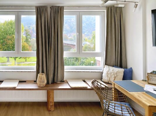MainZauber Apartments früher MainTerrasse Miltenberg am Main - Entdecken Sie die perfekte Luxus-Ferienwohnung mit einem einladenden und hellen Wohnraum. Diese elegante Unterkunft verfügt über große Fenster mit beigen Vorhängen, die einen atemberaubenden Blick auf üppige Bäume und einen malerischen Hügel in der Ferne bieten. Entspannen Sie sich auf einer gemütlichen Holzbank mit Kissen unter diesen malerischen Fenstern oder entspannen Sie sich in einem stilvollen Korbstuhl neben einem wunderschön arrangierten Schreibtisch mit Lampe und geschmackvollem Dekor. Ihre Traumferienwohnung erwartet Sie!