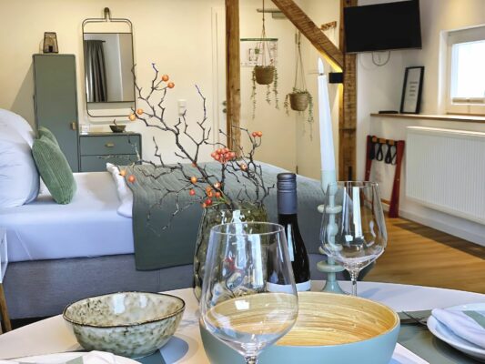MainZauber Apartments früher MainTerrasse Miltenberg am Main - Entdecken Sie mit dieser luxuriösen Unterkunft das ultimative Ferienapartment-Erlebnis. Dieser elegante Raum bietet einen gemütlichen Essbereich mit einem für zwei Personen gedeckten Tisch mit exquisiten Weingläsern und Tellern. Bereichern Sie Ihr Urlaubsessen mit einem auffälligen Zweig-Mittelstück, das mit leuchtend orangefarbenen Beeren geschmückt ist. Das Schlafzimmer des Luxusapartments verfügt über ein bequemes Bett mit stilvollen grünen Kissen sowie eine geschmackvolle Kommode und einen Spiegel für Ihren Komfort. Entspannen Sie sich vor dem an der Wand montierten Fernseher und genießen Sie die entspannende Atmosphäre, die durch durchdacht platzierte Hängepflanzen entsteht, die diesem perfekten Urlaubsdomizil Wärme verleihen.