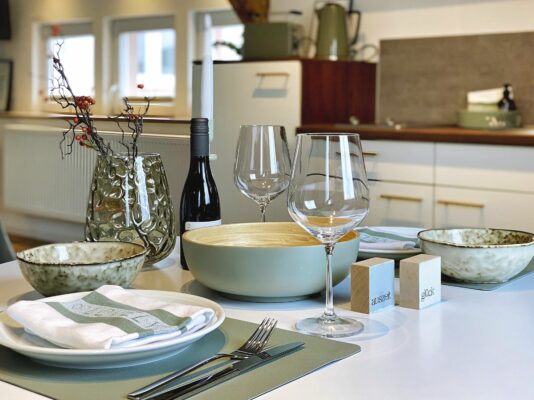 MainZauber Apartments früher MainTerrasse Miltenberg am Main - Betreten Sie Ihr Traumferienapartment und entdecken Sie die perfekte Mischung aus Stil und Komfort. Der offene Essbereich des Luxusapartments verfügt über einen modernen Esstisch, der elegant mit zwei Weingläsern, einer edlen Flasche Wein, Keramikschüsseln, Tellern und Besteck gedeckt ist – ideal für gemütliche Mahlzeiten. Eine schicke grüne Vase mit dekorativen Zweigen und Beeren verleiht dem Raum eine künstlerische Note. Nur wenige Schritte entfernt finden Sie eine voll ausgestattete Küche, die während Ihres Aufenthalts in dieser exquisiten Unterkunft alle Ihre kulinarischen Wünsche erfüllt.