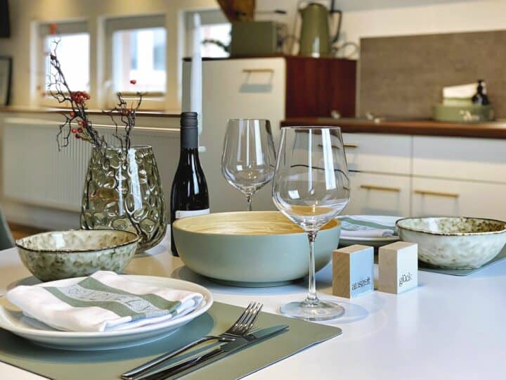 MainZauber Apartments früher MainTerrasse Miltenberg am Main - Betreten Sie Ihr Traumferienapartment und entdecken Sie die perfekte Mischung aus Stil und Komfort. Der offene Essbereich des Luxusapartments verfügt über einen modernen Esstisch, der elegant mit zwei Weingläsern, einer edlen Flasche Wein, Keramikschüsseln, Tellern und Besteck gedeckt ist – ideal für gemütliche Mahlzeiten. Eine schicke grüne Vase mit dekorativen Zweigen und Beeren verleiht dem Raum eine künstlerische Note. Nur wenige Schritte entfernt finden Sie eine voll ausgestattete Küche, die während Ihres Aufenthalts in dieser exquisiten Unterkunft alle Ihre kulinarischen Wünsche erfüllt.