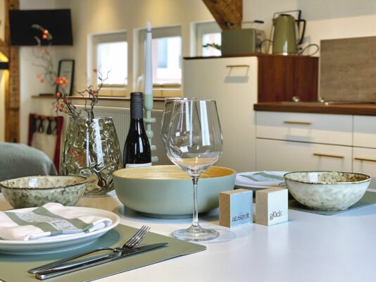 MainZauber Apartments Miltenberg am Main - Erleben Sie den ultimativen Komfort in dieser luxuriösen Ferienwohnung, in der ein gut gedeckter Esstisch die Mahlzeiten in eine elegante Angelegenheit verwandelt. Diese geräumige und gemütliche Wohnküche verfügt über elegante weiße Schränke und stilvolle Holzregale, die eine warme Atmosphäre schaffen, perfekt für Ihre Urlaubsunterkunftsbedürfnisse. Genießen Sie intime Abendessen mit grünen Schüsseln, einem Weinglas und einer Flasche Wein, ergänzt durch geschmackvolle Dekorationselemente. Ihre ideale Ferienwohnung erwartet Sie – buchen Sie jetzt, um sich während Ihres Urlaubs Entspannung und Eleganz zu gönnen.
