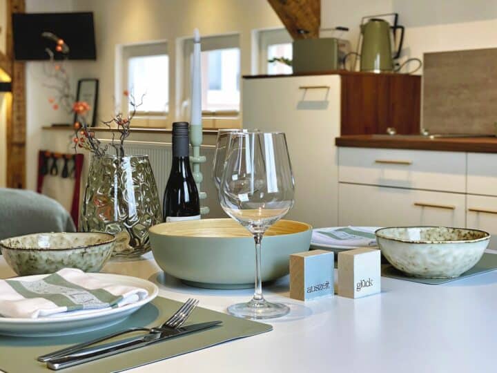 MainZauber Apartments Miltenberg am Main - Erleben Sie den ultimativen Komfort in dieser luxuriösen Ferienwohnung, in der ein gut gedeckter Esstisch die Mahlzeiten in eine elegante Angelegenheit verwandelt. Diese geräumige und gemütliche Wohnküche verfügt über elegante weiße Schränke und stilvolle Holzregale, die eine warme Atmosphäre schaffen, perfekt für Ihre Urlaubsunterkunftsbedürfnisse. Genießen Sie intime Abendessen mit grünen Schüsseln, einem Weinglas und einer Flasche Wein, ergänzt durch geschmackvolle Dekorationselemente. Ihre ideale Ferienwohnung erwartet Sie – buchen Sie jetzt, um sich während Ihres Urlaubs Entspannung und Eleganz zu gönnen.