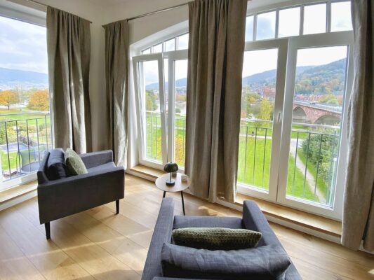 MainZauber Apartments früher MainTerrasse Miltenberg am Main - Entdecken Sie die perfekte Ferienwohnung für Ihren Urlaub! Diese luxuriöse Unterkunft bietet einen hellen und einladenden Raum, hervorgehoben durch große Fenster, die einen atemberaubenden Blick auf eine malerische Brücke und einen ruhigen Fluss zwischen üppigen Bäumen bieten. Im Inneren finden Sie zwei elegante graue Sessel und einen charmanten kleinen runden Tisch, die einen idealen Ort zum Entspannen schaffen. Die hellbraunen Vorhänge rahmen die Aussicht wunderschön ein und verleihen dieser exquisiten Luxuswohnung für den Urlaub Wärme und Stil.