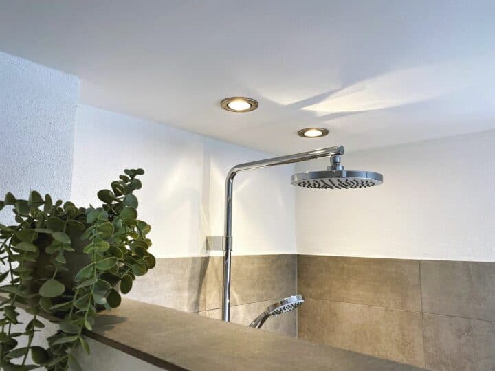 MainZauber Apartments früher MainTerrasse Miltenberg am Main - Entdecken Sie das ultimative Luxusapartment für Ihren Urlaub mit unseren exquisiten Unterkünften, die über ein elegantes, modernes Badezimmer verfügen, das Ihren Aufenthalt noch angenehmer macht. Entspannen Sie sich unter einem elegant an der Wand montierten Regenduschkopf aus Edelstahl. Der Raum wird durch zwei eingelassene Deckenleuchten wunderschön beleuchtet und schafft eine beruhigende Atmosphäre. Für einen zusätzlichen Hauch von Ruhe akzentuiert eine üppige grüne Blattpflanze die Trennwand neben der Dusche. Die Ästhetik des Badezimmers wird durch stilvolle beige-weiße Fliesen vervollständigt, die die Wände schmücken und eine ruhige und erfrischende Atmosphäre während Ihres Urlaubs versprechen.