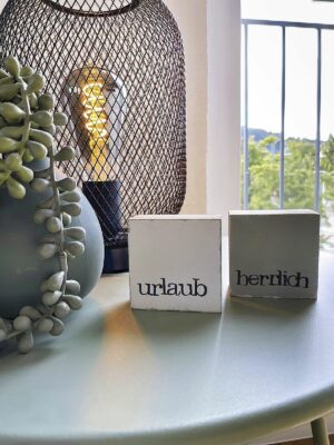 Entdecken Sie die perfekte Ferienwohnung für Ihren nächsten Urlaub! Stellen Sie sich vor, Sie entspannen sich in einem Luxusapartment, umgeben von durchdachten Details, in denen Sie sich wirklich wie zu Hause fühlen. Stellen Sie sich zwei charmante Holzklötze mit der Aufschrift „Urlaub“ und „herzlich“ vor, die einen warmen Ton auf Ihrem gemütlichen, hellblauen Tisch erzeugen. Ihre Unterkunft bietet einzigartige Details wie eine moderne schwarze Netzlampe, die neben einer erfrischenden Sukkulente leuchtet, während ein Fenster mit Blick auf üppiges Grün Ihre ruhige Umgebung bereichert. Buchen Sie dieses außergewöhnliche Luxusapartment noch heute für ein unvergessliches Urlaubserlebnis! - MainZauber Apartments Miltenberg am Main