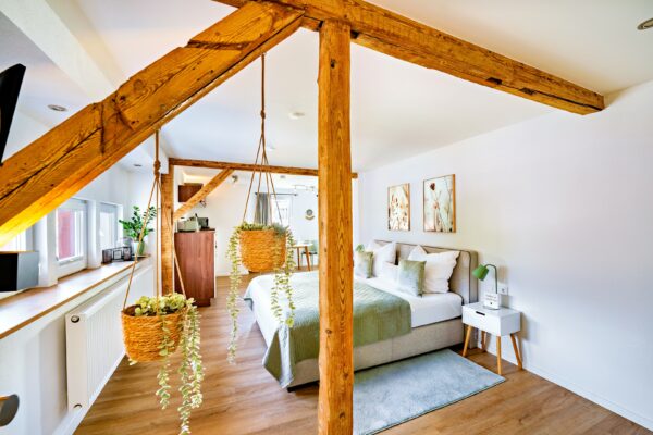 MainZauber Apartments Miltenberg am Main - Entdecken Sie das perfekte Luxusapartment für Ihren Urlaub mit einem charmanten Schlafzimmer, das auf Komfort und Stil ausgelegt ist. Diese einladende Unterkunft bietet eine gemütliche Atmosphäre mit eleganten Holzbalken und einem weichen Bett mit grüner und weißer Bettwäsche. Bereichern Sie Ihren Aufenthalt mit einem Hauch von Natur, den zwei hängende Topfpflanzen verleihen. Die schönen Holzböden des Zimmers ergänzen die sorgfältig kuratierten Kunstwerke an den Wänden, während ein kleiner Nachttisch mit Lampe für zusätzlichen Komfort sorgt. Genießen Sie das natürliche Sonnenlicht, das durch das Fenster fällt, und schaffen Sie so einen idealen Rückzugsort zum Entspannen während Ihres Urlaubs.