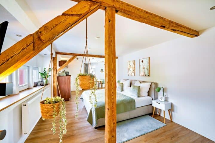 MainZauber Apartments Miltenberg am Main - Entdecken Sie das perfekte Luxusapartment für Ihren Urlaub mit einem charmanten Schlafzimmer, das auf Komfort und Stil ausgelegt ist. Diese einladende Unterkunft bietet eine gemütliche Atmosphäre mit eleganten Holzbalken und einem weichen Bett mit grüner und weißer Bettwäsche. Bereichern Sie Ihren Aufenthalt mit einem Hauch von Natur, den zwei hängende Topfpflanzen verleihen. Die schönen Holzböden des Zimmers ergänzen die sorgfältig kuratierten Kunstwerke an den Wänden, während ein kleiner Nachttisch mit Lampe für zusätzlichen Komfort sorgt. Genießen Sie das natürliche Sonnenlicht, das durch das Fenster fällt, und schaffen Sie so einen idealen Rückzugsort zum Entspannen während Ihres Urlaubs.