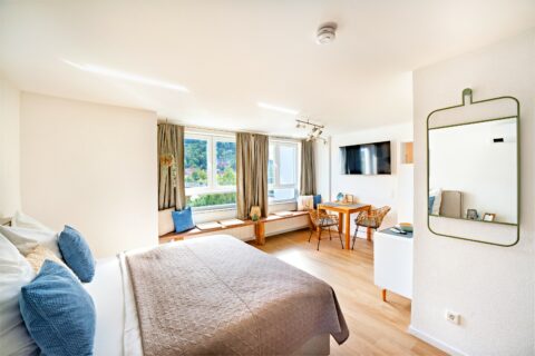 MainZauber Apartments Miltenberg am Main - Entdecken Sie das perfekte Ferienapartment-Erlebnis in dieser luxuriösen Unterkunft. Dieses atemberaubende Luxusapartment für den Urlaub verfügt über ein helles und geräumiges Schlafzimmer mit einem großen Bett, das für Ihren Komfort wunderschön mit blauen und weißen Kissen geschmückt ist. Genießen Sie vom Fenstersitz aus einen atemberaubenden Blick auf die Berge – ein perfekter Ort zum Entspannen nach einem Tag voller Abenteuer. Das Zimmer ist außerdem mit einem an der Wand montierten Fernseher ausgestattet, der Ihnen Unterhaltung auf Knopfdruck bietet. Ein kleiner Schreibtisch mit Stühlen bietet bei Bedarf einen praktischen Arbeitsbereich, während ein zusätzlicher Wandspiegel das moderne und minimalistische Dekor dieses exquisiten Raums ergänzt.