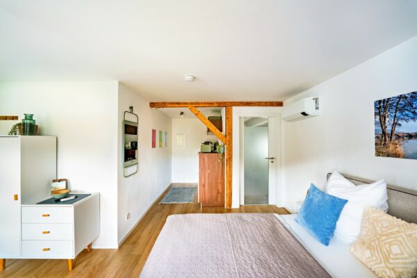 MainZauber Apartments Miltenberg am Main - Entdecken Sie die perfekte Ferienwohnung für Ihren Kurzurlaub – in dieser luxuriösen Unterkunft erwartet Sie ein gemütliches, modernes Schlafzimmer. Genießen Sie ein ordentlich gemachtes Bett mit weichen weißen und blauen Kissen, die für eine erholsame Nachtruhe sorgen. Das Zimmer ist durchdacht eingerichtet mit einer stilvollen Holzkommode und bezaubernder Wandkunst, ergänzt durch einen charmanten kleinen weißen Schrank mit eleganten Dekorationsgegenständen. Ein sichtbarer Holzbalken verleiht diesem hellen und einladenden Raum einen zusätzlichen Hauch von Charme und macht ihn zur idealen Wahl für Ihr nächstes Erlebnis in einer luxuriösen Ferienwohnung.