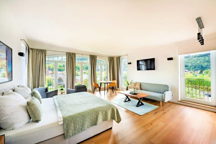 MainZauber Apartments Miltenberg am Main - Erleben Sie das ultimative Leben in einer Ferienwohnung mit unserer Luxusunterkunft. Dieses geräumige, moderne Schlafzimmer verfügt über ein luxuriöses Kingsize-Bett mit eleganter grüner Bettwäsche und einen bequemen Sitzbereich zum Entspannen. Große Fenster bieten einen atemberaubenden Blick auf üppiges Grün und schaffen einen ruhigen Rückzugsort, den Sie lieben werden. Genießen Sie Unterhaltung auf dem an der Wand montierten Fernseher und genießen Sie Mahlzeiten am gemütlichen Esstisch, der bequem am Fenster steht. Buchen Sie noch heute Ihre Luxuswohnung für den Urlaub und gönnen Sie sich einen ruhigen Rückzugsort wie keinen anderen.