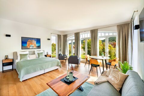 MainZauber Apartments Miltenberg am Main - Entdecken Sie das perfekte Luxus-Ferienapartment-Erlebnis mit diesem geräumigen, gut beleuchteten Schlafzimmer mit einem großen Bett mit stilvoller grüner Bettwäsche und elegantem Holzboden. Diese erstklassige Unterkunft bietet einen gemütlichen Sitzbereich mit plüschigen grauen Stühlen und einem schicken runden Esstisch, alles eingerahmt von großen Fenstern mit hübschen Vorhängen, die einen malerischen Blick auf die Bäume draußen bieten. Zeitgenössisches Dekor wertet jede Ecke dieses exquisiten Ferienapartments auf und sorgt für einen unvergesslichen Aufenthalt.