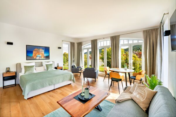 MainZauber Apartments Miltenberg am Main - Entdecken Sie das perfekte Luxus-Ferienapartment-Erlebnis mit diesem geräumigen, gut beleuchteten Schlafzimmer mit einem großen Bett mit stilvoller grüner Bettwäsche und elegantem Holzboden. Diese erstklassige Unterkunft bietet einen gemütlichen Sitzbereich mit plüschigen grauen Stühlen und einem schicken runden Esstisch, alles eingerahmt von großen Fenstern mit hübschen Vorhängen, die einen malerischen Blick auf die Bäume draußen bieten. Zeitgenössisches Dekor wertet jede Ecke dieses exquisiten Ferienapartments auf und sorgt für einen unvergesslichen Aufenthalt.