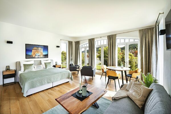 Entdecken Sie Ihre ideale Urlaubsunterkunft in unserem Luxusapartment mit einem geräumigen und modernen Schlafzimmer, das für Ihren perfekten Urlaub konzipiert wurde. Entspannen Sie sich auf dem großen, bequemen Bett, umgeben von eleganten Holzböden. Genießen Sie eine gemütliche Sitzecke mit einem Plüschsofa, einem stilvollen Tisch und einladenden Sesseln. Das Zimmer ist durch große, mit Vorhängen geschmückte Fenster in natürliches Licht getaucht, das einen ruhigen Blick auf das üppige Grün draußen freigibt. Werten Sie Ihren Aufenthalt mit geschmackvoller Einrichtung auf, darunter lebendige Pflanzen und faszinierende Wandkunst. Buchen Sie noch heute Ihr Traumferienapartment für ein unvergessliches Erlebnis! - MainZauber Apartments Miltenberg am Main