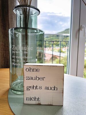 MainZauber Apartments früher MainTerrasse Miltenberg am Main - Entdecken Sie Ihre Traumferienwohnung im Herzen von Miltenberg, wo Luxus auf atemberaubende Landschaft trifft. Stellen Sie sich vor, Sie entspannen in einer luxuriösen Unterkunft mit einem atemberaubenden Fensterblick auf grüne Hügel und einen magischen Wolkenhimmel. Auf dem Holztisch trägt eine bezaubernde Glasflasche zur Atmosphäre bei, während ein Holzblock den bezaubernden deutschen Spruch „ohne Zauber geht’s auch nicht“ trägt. Tauchen Sie ein in diese idyllische MainZauber-Umgebung und schaffen Sie unvergessliche Erinnerungen in Ihrer perfekten Luxuswohnung für den Urlaub.