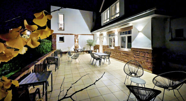 MainZauber Apartments früher MainTerrasse Miltenberg am Main - Entdecken Sie den perfekten Rückzugsort in unserer luxuriösen Ferienwohnung mit einer wunderschön beleuchteten Außenterrasse, die ideal zum Entspannen ist. Entspannen Sie stilvoll in gemütlichen Sitzgelegenheiten mit bequemen Tischen und Stühlen inmitten üppiger Vegetation. Das elegante, moderne Gebäude verfügt über große Fenster, die Ihr Wohnerlebnis mit reichlich natürlichem Licht bereichern. Leuchtend gelbe Blumen verleihen dieser ruhigen Oase einen lebendigen Farbtupfer und machen sie zu einer idyllischen Kulisse für Ihren nächsten Urlaub.