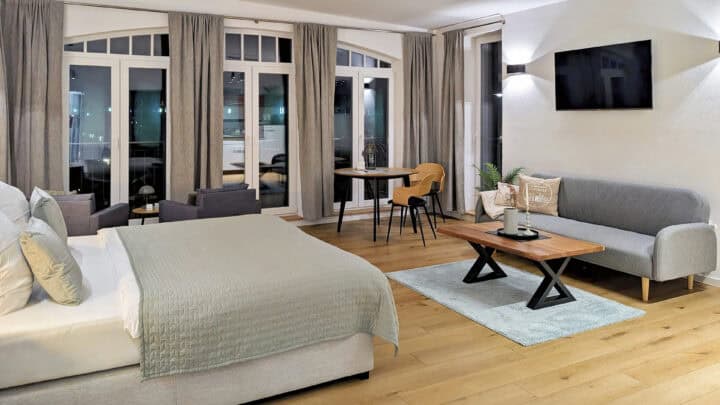 MainZauber Apartments früher MainTerrasse Miltenberg am Main - Entdecken Sie die perfekte Ferienwohnung, die moderne Eleganz und Komfort für Ihren Urlaub vereint. Diese luxuriöse Unterkunft verfügt über ein geräumiges Schlafzimmer mit einem großen, einladenden Bett mit stilvoller grauer Bettwäsche. Der Holzboden unterstreicht das anspruchsvolle Ambiente, während eine gemütliche Sitzecke zum Entspannen einlädt. Genießen Sie Mahlzeiten am gemütlichen Esstisch mit Stühlen und entspannen Sie sich vor dem an der Wand montierten Fernseher. Die Wohnung verfügt über große Fenster mit edlen grauen Vorhängen, die den Raum mit natürlichem Licht füllen und seine neutralen Töne für eine warme und einladende Atmosphäre beibehalten. Buchen Sie diese exquisite Luxuswohnung noch heute für Ihren nächsten Urlaub!