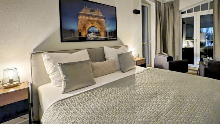 MainZauber Apartments früher MainTerrasse Miltenberg am Main - Erleben Sie in dieser luxuriösen Ferienwohnung das Höchstmaß an Komfort und Stil. Das gemütliche Schlafzimmer verfügt über ein geräumiges, weiches Bett mit eleganten weißen und grauen Kissen, ergänzt durch eine stilvolle strukturierte graue Tagesdecke. Moderne Lampen auf schicken Nachttischen aus Holz sorgen für ein stimmungsvolles Licht für einen erholsamen Rückzugsort. Ein wunderschön gerahmtes Bild eines beleuchteten Bogens bei Nacht verleiht der Einrichtung des Zimmers einen Hauch kunstvoller Eleganz. Vom Boden bis zur Decke reichende Vorhänge verdecken elegant die Glastüren, die auf Ihren eigenen Balkon oder Ihre Terrasse führen – ein perfekter Ort zum Entspannen und Aufsaugen der ruhigen Umgebung Ihrer Unterkunft. Ideal für alle, die für ihren nächsten Urlaub eine gehobene Unterkunft suchen!