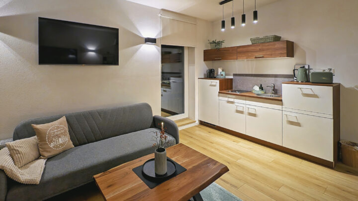 MainZauber Apartments früher MainTerrasse Miltenberg am Main - Erleben Sie den ultimativen Komfort in einer Ferienwohnung mit unserer luxuriösen Unterkunft. Dieser elegante Raum verfügt über einen gemütlichen, modernen Wohn- und Küchenbereich, perfekt für Ihren Kurzurlaub. Entspannen Sie auf dem stilvollen, grauen Sofa mit den weichen Kissen, während Sie Unterhaltung auf dem an der Wand montierten Fernseher genießen. Der Wohnbereich umfasst auch einen charmanten Couchtisch aus Holz, der ein anspruchsvolles und dennoch einladendes Ambiente schafft.

Unsere Luxuswohnung verfügt über eine kompakte Küche mit schönen Holzschränken, ideal zum Zubereiten von Mahlzeiten während Ihres Aufenthalts. Sanfte Beleuchtung sorgt überall für eine warme und einladende Atmosphäre und verbessert Ihr Entspannungserlebnis. Eine Tür führt bequem in ein weiteres Zimmer und bietet Ihnen zusätzlichen Platz zum Entspannen während Ihres Traumurlaubs in diesem exquisiten Rückzugsort. Entdecken Sie noch heute unvergleichliche Gemütlichkeit und Stil in unserer erstklassigen Ferienunterkunft!