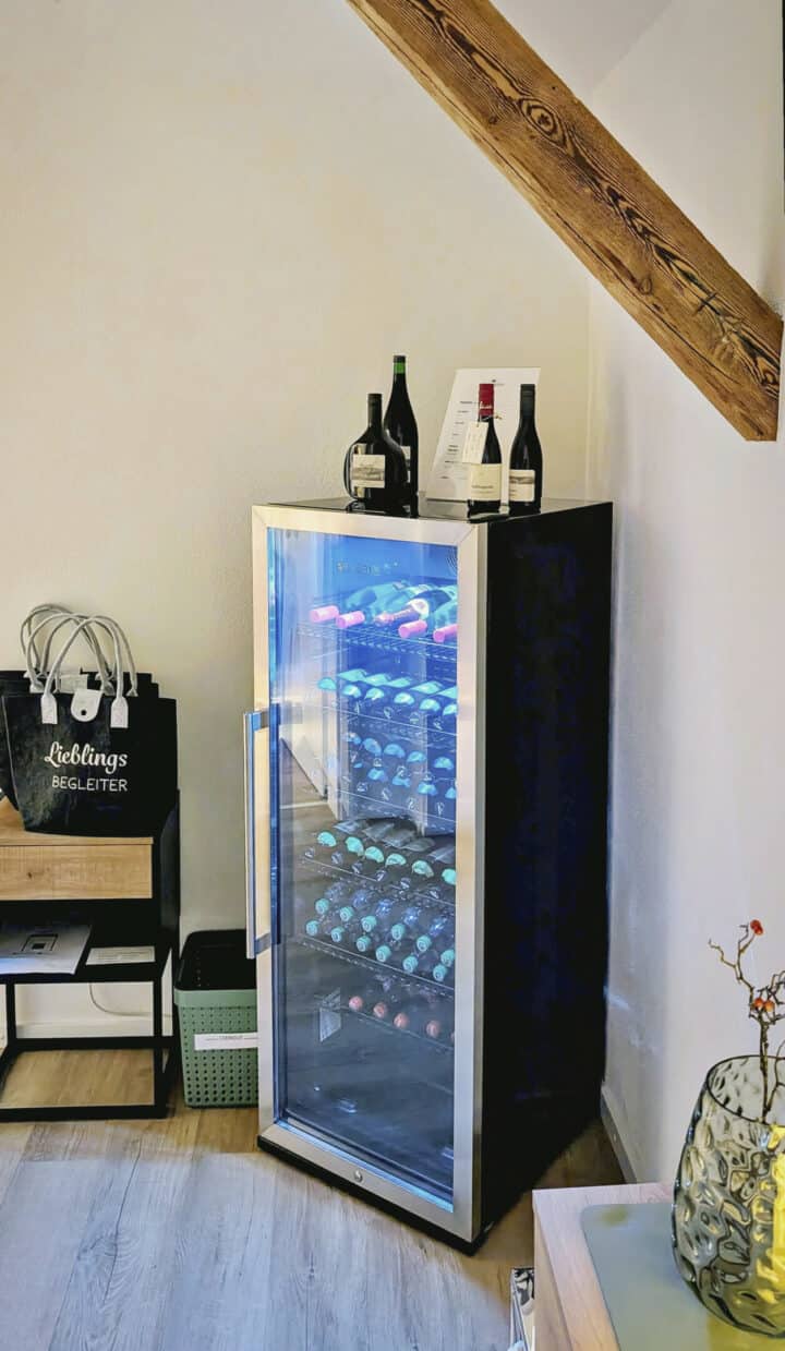 MainZauber Apartments früher MainTerrasse Miltenberg am Main - Entdecken Sie die Ferienwohnung Ihrer Träume, in der Luxus auf Komfort trifft. In der Ecke unserer großzügigen Unterkunft steht ein eleganter schwarzer Weinkühler, der elegant mit erlesenen Flaschen für Ihren Genuss gefüllt ist. Dieser Kühler steht unter einem bezaubernden Holzbalken und verfügt über eine elegante Glastür, auf der drei Premiumflaschen ausgestellt sind, was das edle Ambiente des Raums noch verstärkt. In den Regalen daneben sind schicke Tragetaschen und stilvolle Dekorationsartikel ausgestellt, die dafür sorgen, dass Ihr Aufenthalt in unserem Luxusapartment für den Urlaub nichts weniger als außergewöhnlich wird. Gönnen Sie sich diesen perfekten Kurzurlaub und steigern Sie Ihr Urlaubserlebnis bei uns!