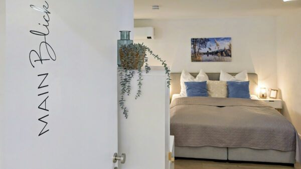 MainZauber Apartments früher MainTerrasse Miltenberg am Main - Entdecken Sie Ihre ideale Ferienwohnung in unserer luxuriösen Unterkunft. Unser elegant gestaltetes Schlafzimmer verfügt über ein ordentlich gemachtes Bett mit weichen grauen und blauen Kissen, das Ihnen während Ihres Aufenthalts den ultimativen Komfort bietet. Über dem Bett hängt ein bezauberndes Landschaftsgemälde, das zur ruhigen Atmosphäre dieser luxuriösen Ferienwohnung beiträgt. Wenn Sie durch die weiße Tür mit der Aufschrift „Main Blick“ treten, finden Sie eine stilvolle Einrichtung, darunter eine bezaubernde Zierpflanze – ein perfekter Rückzugsort für Ihren Traumurlaub.
