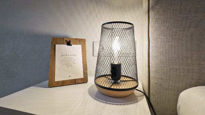 MainZauber Apartments früher MainTerrasse Miltenberg am Main - Erleben Sie die perfekte Mischung aus Stil und Komfort in Ihrer Ferienwohnung. Diese luxuriöse Ferienwohnung verfügt über eine moderne Nachttischlampe mit einem schicken Schirm aus Drahtgeflecht, die ein warmes Licht in den Raum bringt. Neben der Lampe befindet sich ein wunderschön gearbeiteter Holzrahmen mit einem eleganten Zitat, der auf einem makellos weißen Nachttisch ruht und eine einladende und anspruchsvolle Atmosphäre für Ihren Urlaubsort schafft.