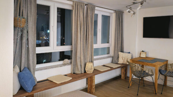 MainZauber Apartments früher MainTerrasse Miltenberg am Main - Entdecken Sie die perfekte Ferienwohnung, in der Komfort auf Stil trifft. Diese luxuriöse Unterkunft verfügt über einen gemütlichen Wohnbereich mit einer charmanten Holzbank unter großen Fenstern, die mit weichen Kissen geschmückt sind – Ihr idealer Ort zum Entspannen. Genießen Sie Ihre Mahlzeiten in der schicken Essecke mit einem stilvollen kleinen Tisch und modernen Drahtstühlen. Mit einem an der Wand montierten Fernseher und modernem Dekor in neutralen Tönen bietet dieses erstklassige Luxusapartment eine einladende Atmosphäre für Ihren Traumurlaub.