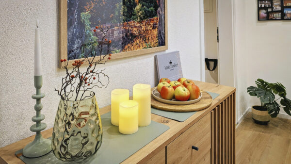 MainZauber Apartments früher MainTerrasse Miltenberg am Main - Entdecken Sie das perfekte Ferienapartment-Erlebnis mit unserer luxuriösen Unterkunft. Dieser elegant gestaltete Raum verfügt über ein gemütliches Interieur, komplett mit einem hölzernen Sideboard, auf dem dekorative Kerzen wunderschön zur Geltung kommen, einer stilvollen Vase mit Zweigen und leuchtend roten Beeren und einer Obstschale voller frischer Äpfel. Verschönern Sie Ihren Aufenthalt in unserem luxuriösen Urlaubsapartment, indem Sie die bezaubernde gerahmte Herbstlandschaftskunst bewundern, die die weiße Wand ziert, während eine bezaubernde Topfpflanze dem einladenden Ambiente mit Holzboden Frische verleiht.