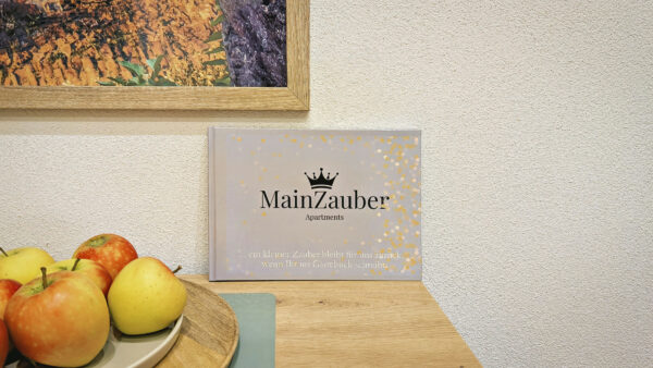 MainZauber Apartments früher MainTerrasse Miltenberg am Main - Entdecken Sie Ihren perfekten Urlaub mit „MainZauber Apartments“, prominent präsentiert auf einem Tisch an einer Wand. Dieses Luxusapartment für Urlaubssuchende verspricht außergewöhnliche Unterkunftserlebnisse. Eine Schale mit frischen Äpfeln sorgt für eine einladende Note, während der Buchumschlag, geschmückt mit einem eleganten Kronendesign und schimmernden, goldenen, konfettiartigen Mustern, auf den Luxus hinweist, der Sie erwartet. Werten Sie Ihren Urlaubsaufenthalt auf, indem Sie Unterkünfte wählen, die sowohl Stil als auch Komfort bieten. Ein darüber hängendes gerahmtes Bild vervollständigt diese einladende Aussicht und macht deutlich, dass „MainZauber Apartments“ der Ort ist, an dem jedes Element Ihres Urlaubs sorgfältig zusammengestellt wird, um unvergessliche Erinnerungen zu schaffen.