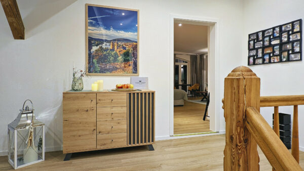 MainZauber Apartments früher MainTerrasse Miltenberg am Main - Treten Sie in den einladenden Flur Ihrer Traum-*Ferienwohnung*, wo Sie ein charmantes Sideboard aus Holz mit einer Reihe von Kerzen und einer Schale mit frischem Obst begrüßt und eine Atmosphäre der Wärme und des Willkommens schafft. Über dem Sideboard bietet ein atemberaubendes Landschaftsgemälde einen Blick auf ruhige Aussichten. Eine stilvolle Laterne trägt zur gemütlichen Atmosphäre bei, während Sie auf der rechten Seite eine Tür einlädt, einen gut beleuchteten Raum zu erkunden, der mit elegant gerahmten Fotos geschmückt ist. Dieses *Luxus-Apartment für den Urlaub* verspricht nicht nur eine außergewöhnliche Unterkunft, sondern auch einen unvergesslichen Aufenthalt, der Komfort mit Stil verbindet.