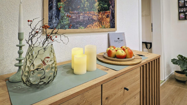 MainZauber Apartments früher MainTerrasse Miltenberg am Main - Entdecken Sie die perfekte Ferienwohnung, die Komfort und Eleganz vereint. Stellen Sie sich vor, Sie befinden sich in einem wunderschön dekorierten Raum mit einem luxuriösen Sideboard aus Holz, das durch eine dekorative Glasvase mit Zweigen und roten Beeren noch aufgewertet wird. Entspannen Sie sich neben zwei brennenden Kerzen, die einen warmen Schein über den Raum werfen, und genießen Sie dazu einen Teller mit frischen Äpfeln. Ein gerahmtes Foto verleiht eine persönliche Note, während üppiges Grün in der Ecke das gemütliche Ambiente verstärkt. Diese exquisite Unterkunft verspricht ein unvergessliches Erlebnis für Ihren nächsten Urlaub.