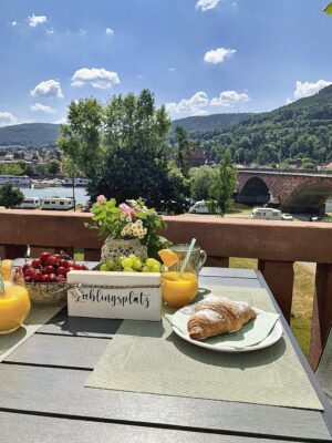 MainZauber Apartments Miltenberg am Main - Stellen Sie sich vor, Sie beginnen Ihren Tag in einer luxuriösen Ferienwohnung mit einem malerischen Frühstück auf dem Balkon. Mit Blick auf einen ruhigen Fluss und eine charmante Brücke bietet diese Unterkunft eine idyllische Umgebung zum Entspannen. Genießen Sie frische Croissants, saftige Kirschen und Trauben, leuchtende Blumen und erfrischende Gläser Orangensaft – und das alles mit dem üppigen, bewaldeten Hügel, der eine wunderschöne Kulisse unter einem wunderschönen blauen Himmel mit vereinzelten Wolken bildet. Machen Sie diese traumhafte Aussicht zu einem Teil Ihres perfekten Urlaubs in unseren exklusiven Ferienwohnungen.