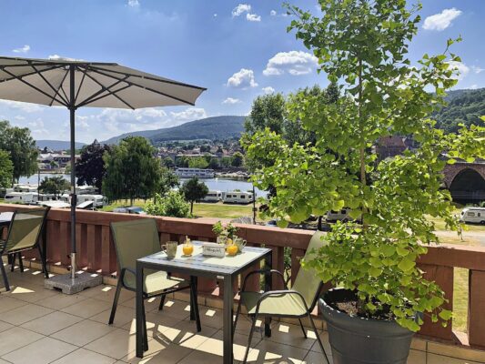 MainZauber Apartments Miltenberg am Main - Erleben Sie die ultimative Urlaubsunterkunft mit unserem Luxusapartment mit sonniger Terrasse und atemberaubender Aussicht. Dieser Außenbereich ist perfekt für alle, die einen erstklassigen Urlaub verbringen möchten. Er ist mit einem Tisch und Stühlen ausgestattet und eine große Topfpflanze verleiht dem Ganzen eine besondere Atmosphäre. Entspannen Sie sich bequem im Schatten eines Sonnenschirms und genießen Sie die malerische Landschaft. Sie werden von den in der Ferne geparkten Wohnmobilen und sanften Hügeln vor dem Hintergrund des blauen Himmels mit flauschigen Wolken fasziniert sein. Das ist Urlaubsleben vom Feinsten!