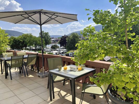 MainZauber Apartments Miltenberg am Main - Entdecken Sie eine atemberaubende Luxus-Ferienwohnung mit sonnendurchfluteter Außenterrasse, perfekt für Ihren Urlaub. Diese exquisite Unterkunft verfügt über mehrere Tische und Stühle unter großen Sonnenschirmen und bietet eine ruhige Umgebung zum Entspannen oder für Unterhaltung. Genießen Sie erfrischende Getränke und genießen Sie dabei die atemberaubende Aussicht auf die üppige Landschaft, die sanften Hügel und den malerischen blauen Himmel mit seinen Wolken. Erleben Sie unvergleichlichen Komfort und Stil in dieser außergewöhnlichen Ferienwohnung.