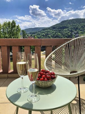 MainZauber Apartments Miltenberg am Main - Stellen Sie sich vor, Sie entspannen sich in einer luxuriösen Ferienwohnung, wo auf dem grünen Tisch des Balkons zwei Gläser Champagner und eine Schale Kirschen auf Sie warten. Entspannen Sie sich im weißen Stuhl und genießen Sie die atemberaubende Aussicht auf üppige grüne Hügel unter einem malerischen Wolkenhimmel. Diese erstklassige Unterkunft verspricht ein unvergessliches Urlaubserlebnis.