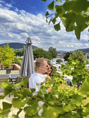 MainZauber Apartments Miltenberg am Main - Ein lächelndes Paar mit Sonnenbrille genießt einen sonnigen Tag in seiner luxuriösen Ferienwohnung. Sie stehen auf einem privaten Balkon, umgeben von üppigem Grün und hoch aufragenden grünen Blättern, mit atemberaubender Aussicht auf ferne Hügel unter einem teilweise bewölkten Himmel. Die perfekte Luxusunterkunft für Ihren nächsten Urlaub, komplett mit Sitzbereichen im Freien und Tischen mit Sonnenschirmen für ultimative Entspannung inmitten der Schönheit der Natur.