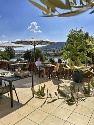 MainZauber Apartments Miltenberg am Main - Stellen Sie sich vor, Sie entspannen sich in einer Ferienwohnung und genießen einen entspannten Morgen auf der sonnigen Außenterrasse. Diese luxuriöse Unterkunft verfügt über Sonnenschirme und Topfpflanzen und schafft so eine einladende Atmosphäre für Ihren Urlaub. Die Aussicht bietet einen atemberaubenden Blick auf einen malerischen Fluss und üppige Hügel unter einem klaren blauen Himmel mit flauschigen Wolken. In der Nähe sind charmante Tische elegant mit Tabletts und Blumen gedeckt und unterstreichen die ruhige Atmosphäre Ihres perfekten Urlaubsdomizils.
