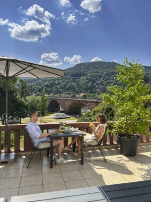 MainZauber Apartments Miltenberg am Main - Stellen Sie sich vor, Sie faulenzen auf der sonnigen Terrasse einer luxuriösen Ferienwohnung, wo die perfekte Unterkunft für Ihren Urlaub auf Sie wartet. Stellen Sie sich vor, Sie schlürfen erfrischende Getränke und genießen köstliche Snacks, während Sie die atemberaubende Aussicht auf eine bezaubernde Steinbrücke genießen, umgeben von üppigen grünen Hügeln unter einem klaren blauen Himmel. Der sanfte Schatten eines Sonnenschirms sorgt für Ihren Komfort, während Sie in dieser idyllischen Umgebung entspannen. Entdecken Sie noch heute die Ferienwohnung Ihrer Träume und erleben Sie unvergleichliche Ruhe und Eleganz.