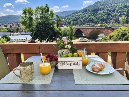 MainZauber Apartments Miltenberg am Main - Gönnen Sie sich ein luxuriöses Ferienapartment-Erlebnis mit einem malerischen Frühstücksplatz im Freien, der einen atemberaubenden Blick auf den Fluss und die Brücke bietet. Diese perfekte Unterkunft ermöglicht es Ihnen, Ihren Morgenkaffee in ruhiger Umgebung zu genießen. Der Tisch ist elegant mit köstlichen Croissants, frischen Kirschen, erfrischendem Orangensaft und bezaubernden Blumen neben einem entzückenden Schild mit der Aufschrift „Lieblingsplatz“ geschmückt. Tauchen Sie ein in die Ruhe der üppigen grünen Hügel und Bäume, die diese idyllische Urlaubsatmosphäre vervollständigen. Reservieren Sie noch heute Ihr Luxusapartment für den Urlaub und machen Sie jeden Morgen zu einer wertvollen Erinnerung.