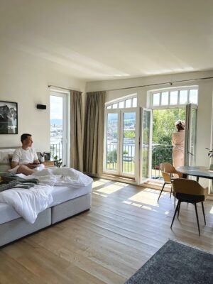Erleben Sie den ultimativen Kurzurlaub mit dieser atemberaubenden Ferienwohnung, perfekt für alle, die eine luxuriöse Wohnung suchen. Betreten Sie das helle Schlafzimmer mit großen Fenstern und einem privaten Balkon, der einen atemberaubenden Blick auf einen malerischen Fluss bietet. Stellen Sie sich vor, Sie entspannen sich mit Ihrem Telefon auf dem bequemen Bett oder treten auf den Balkon, um die ruhige Umgebung zu genießen. Das Zimmer ist elegant eingerichtet, komplett mit einem gemütlichen Bett, einem praktischen Tisch und Stühlen sowie gerahmten Kunstwerken, die Ihrem Aufenthalt einen Hauch von Raffinesse verleihen. Entdecken Sie Urlaubsleben vom Feinsten – ideal, um während Ihres Urlaubs unvergessliche Erinnerungen zu schaffen. - MainZauber Apartments Miltenberg am Main