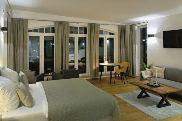 MainZauber Apartments früher MainTerrasse Miltenberg am Main - Entdecken Sie eine luxuriöse Ferienwohnung, die perfekt für Ihren nächsten Urlaub ist. Diese moderne Unterkunft bietet einen geräumigen Wohnbereich mit Holzböden und einem stilvollen Rundfenster mit eleganten grauen Vorhängen. Entspannen Sie sich auf dem plüschigen grauen Sofa, während Sie Ihre Lieblingssendungen auf dem an der Wand montierten Fernseher genießen, oder genießen Sie Mahlzeiten im gut ausgestatteten Essbereich mit Sitzgelegenheiten für zwei Personen am Fenster. Verwöhnen Sie sich mit Komfort und Eleganz in dieser atemberaubenden Luxus-Ferienwohnung, die anspruchsvollen Reisenden ein außergewöhnliches Erlebnis bieten soll.