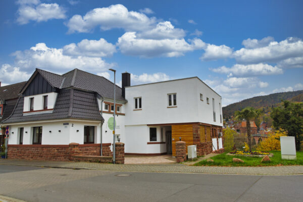 MainZauber Apartments früher MainTerrasse Miltenberg am Main - Entdecken Sie Ihre Traum-**Ferienwohnung** in diesem atemberaubenden modernen weißen Haus mit eleganten Holzakzenten und einem raffinierten dunklen Dach. Dieses **Luxus-Ferienapartment** liegt ideal an einer charmanten Vorstadtstraßenecke und bietet alles, was Sie für einen perfekten Urlaub brauchen. Umgeben von einem gepflegten Rasen bietet das Anwesen einen ruhigen Blick auf einen Kirchturm und malerische Häuser mit roten Dächern unter einem teilweise bewölkten blauen Himmel. Erleben Sie das Höchstmaß an Entspannung und Komfort mit dieser exklusiven Unterkunftsoption, ideal für Reisende, die während ihres Aufenthalts sowohl Luxus als auch Ruhe suchen.