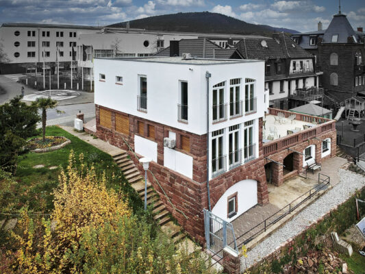 MainZauber Apartments früher MainTerrasse Miltenberg am Main - Entdecken Sie Ihre perfekte Ferienwohnung in dieser modernen Luxusunterkunft. Dieses makellos weiße Gebäude liegt an einem wunderschön angelegten Hang und verfügt über große Fenster, die natürliches Licht hereinlassen. Die charmante untere Holzfassade verleiht einen Hauch von Eleganz, während die geräumige Terrasse Tische und Stühle bietet, damit Sie sich entspannen und die Aussicht genießen können. Mit den umliegenden Gebäuden und einem malerischen Hügel im Hintergrund verspricht diese außergewöhnliche Luxus-Ferienwohnung einen unvergesslichen Aufenthalt unter einem malerischen Wolkenhimmel.