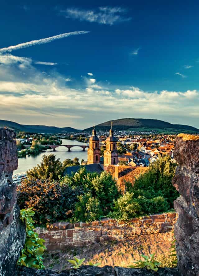 MainZauber Apartments früher MainTerrasse Miltenberg am Main - Entdecken Sie das perfekte Luxusapartment für Ihren Urlaub mit atemberaubender Aussicht auf eine bezaubernde Stadtlandschaft. Wachen Sie mit einem faszinierenden Anblick auf zwei ikonische Kirchtürme auf, die sich elegant inmitten üppiger Vegetation erheben, alles wunderschön eingerahmt von einem ruhigen Fluss, der unter einer historischen Steinbrücke hindurchfließt. Während Sie in dieser exquisiten Unterkunft entspannen, genießen Sie die atemberaubende Kulisse sanfter Hügel unter einem teilweise bewölkten blauen Himmel. Erleben Sie unvergleichlichen Komfort und Eleganz in diesem idealen Ferienapartment, das einen unvergesslichen Urlaub verspricht.